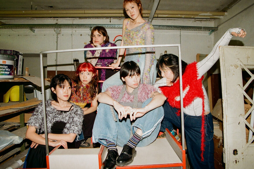 Die sechs koreanischen Künstlerinnen des Punk-Performance-Kollektivs Horizontaler Gentransfer sietzen in einem Atelier auf einem Tisch und präsentieren sich der Kamera. Im Hintergrund sieht man Regale mit Malerutensilien.